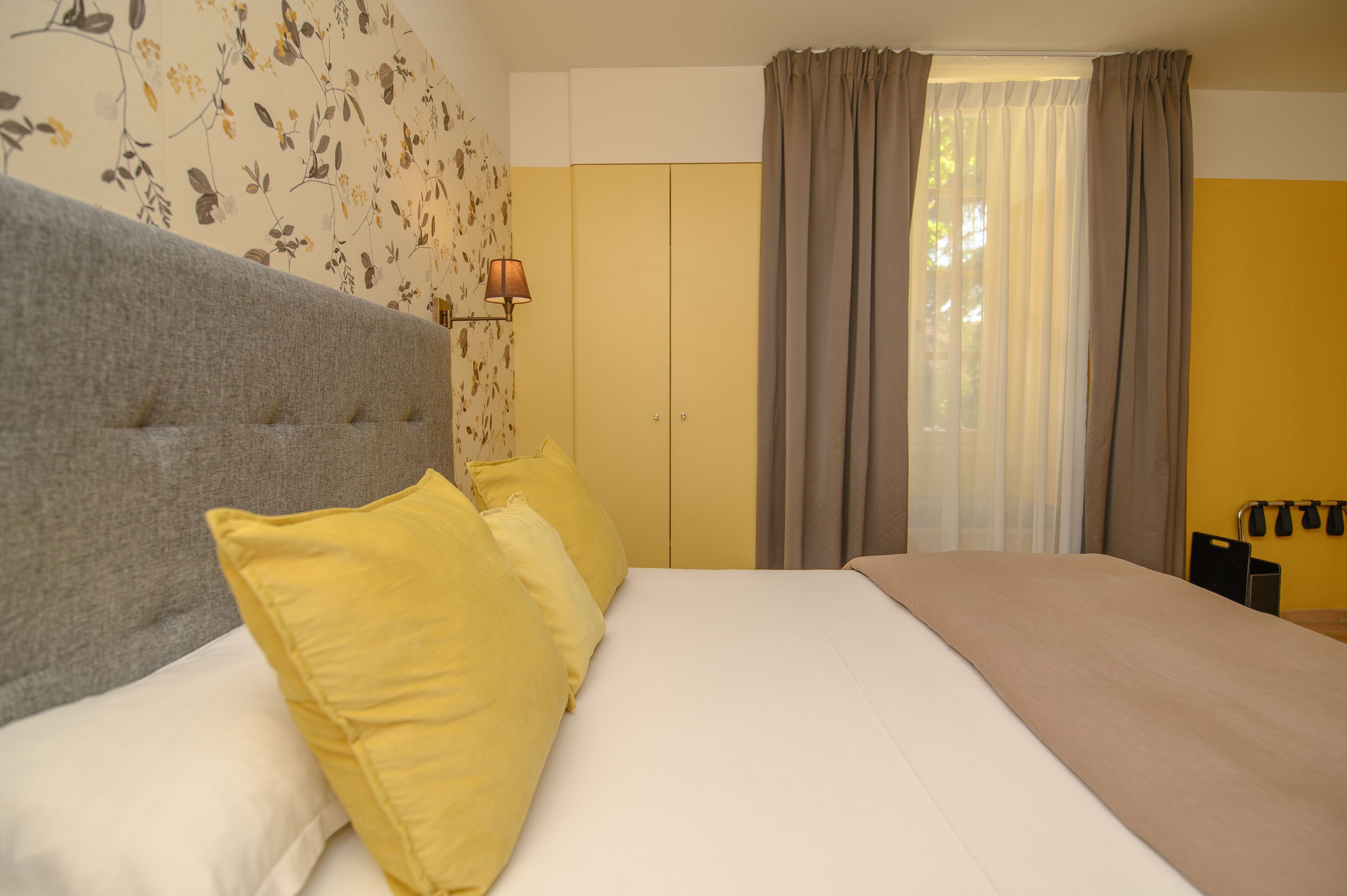 Un dormitorio con una cama grande, dos almohadas, cabecero, pared con papel tapiz floral, ventana con cortinas y una lámpara de pared.