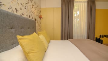 Un dormitorio con una cama grande, dos almohadas, cabecero, pared con papel tapiz floral, ventana con cortinas y una lámpara de pared.