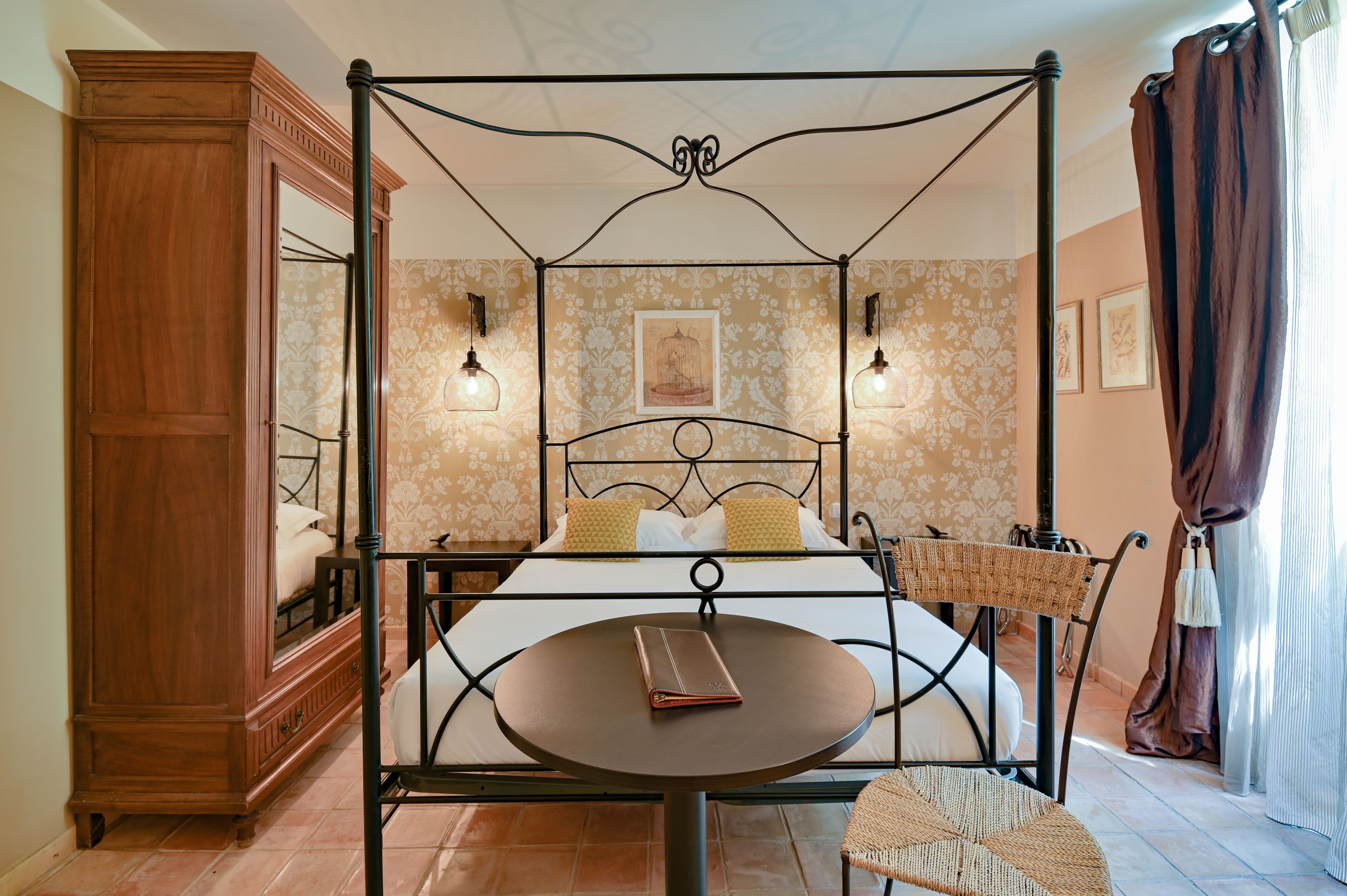 A four-poster bed with a round table in front, a wooden cabinet, and a mirror.