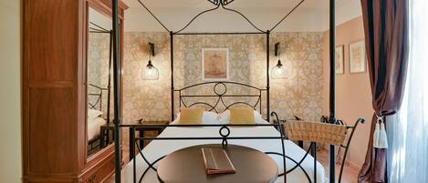 A four-poster bed with a round table in front, a wooden cabinet, and a mirror.