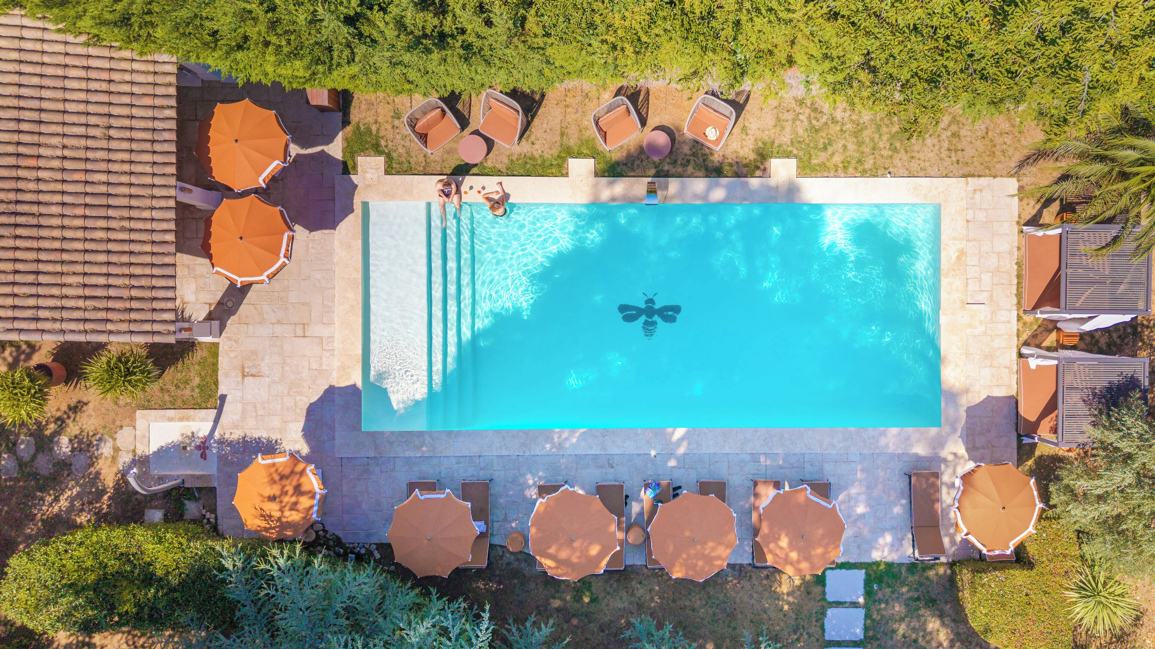 An aerial view of a swimming pool with lounge chairs and umbrellas, surrounded by greenery and a tiled roof.