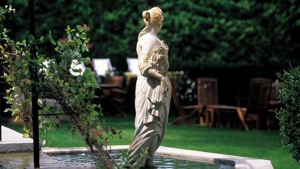 A statue in a garden with a fountain, surrounded by greenery and outdoor furniture. - Villa Augusta (Saint-Paul-Trois-Chateaux)