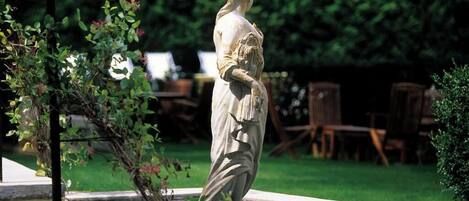 Una estatua en un jardín con una fuente, rodeada de vegetación y mobiliario de exterior.
