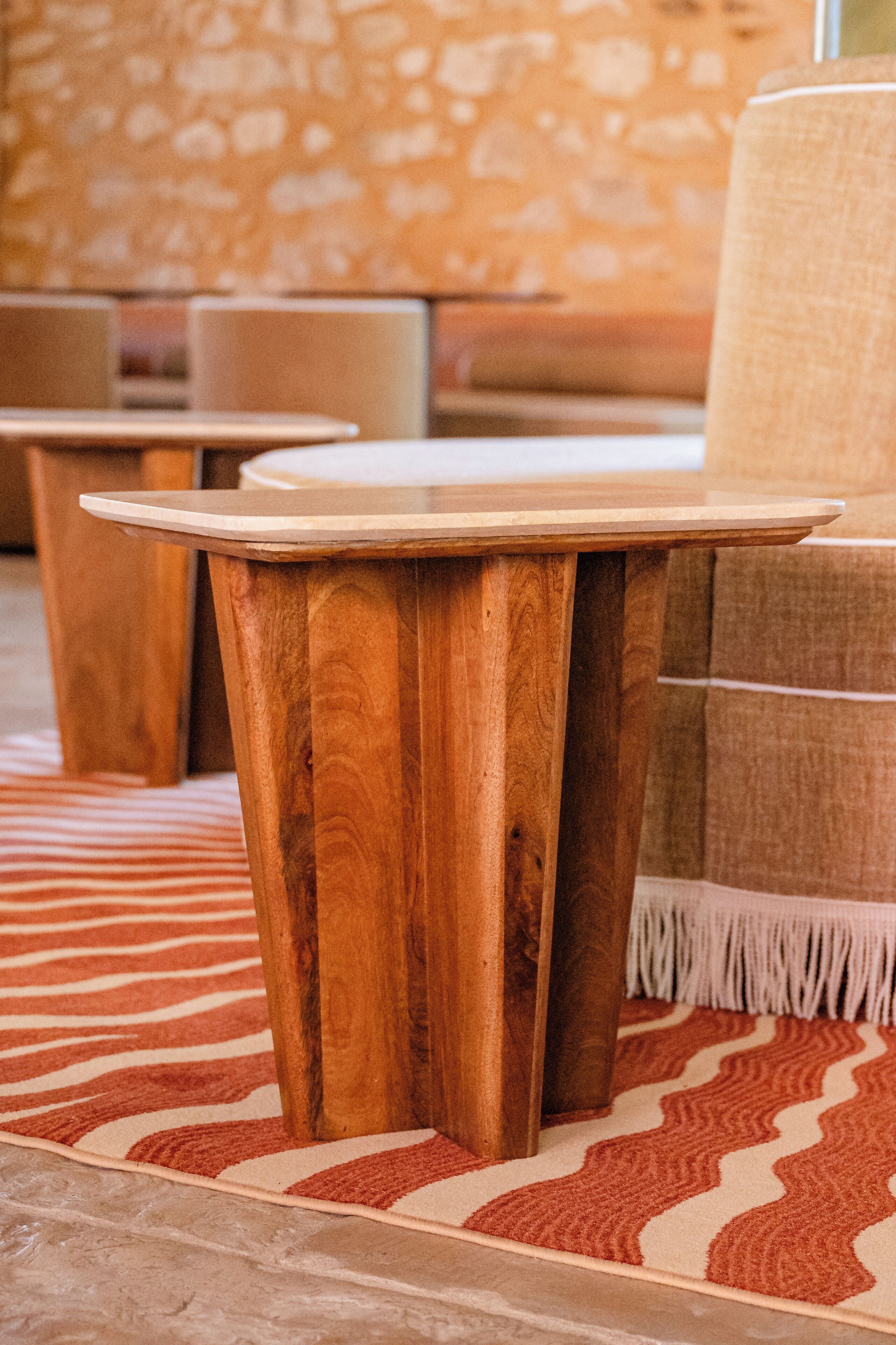 A wooden table with a round top and tapered legs, placed on a patterned rug in a room with stone walls and a cushioned seating area.