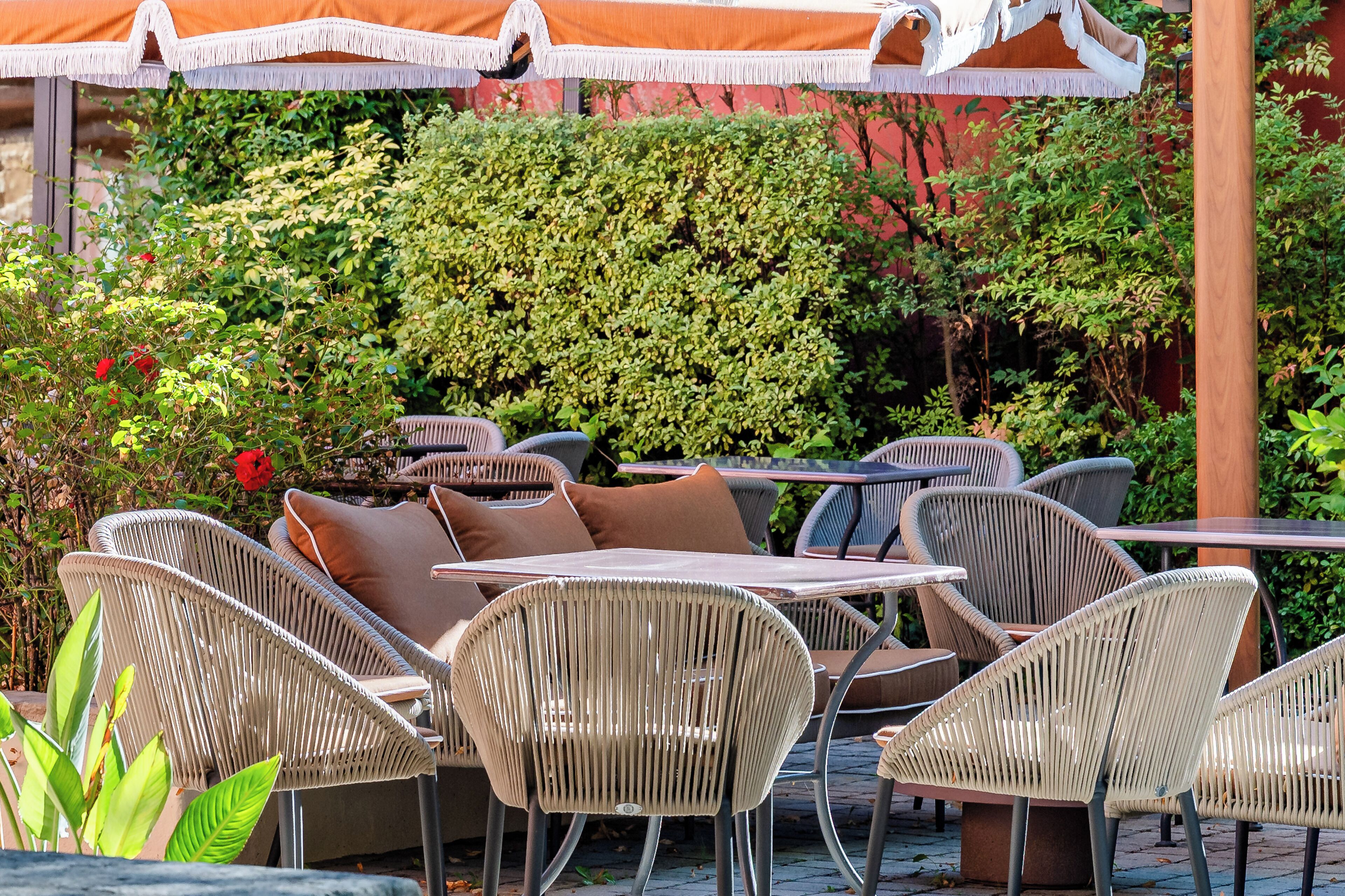 An outdoor seating area with wicker chairs and tables, surrounded by lush greenery and a red wall.