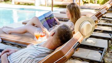 A man lying on a sun lounger by a pool, reading a book.