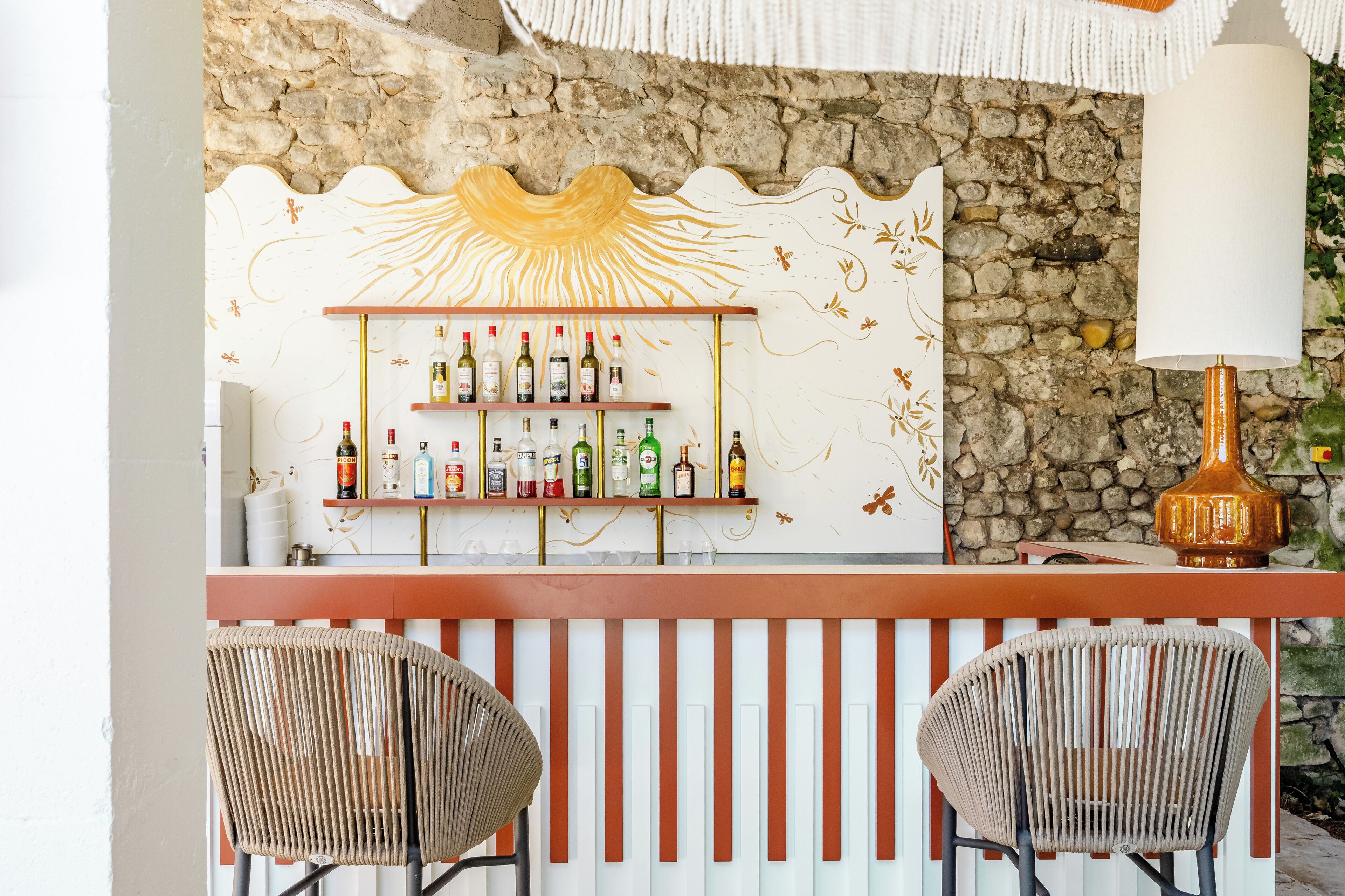 Un coin bar avec un mur en pierre en arrière-plan, une étagère remplie de différentes bouteilles et deux fauteuils en osier.