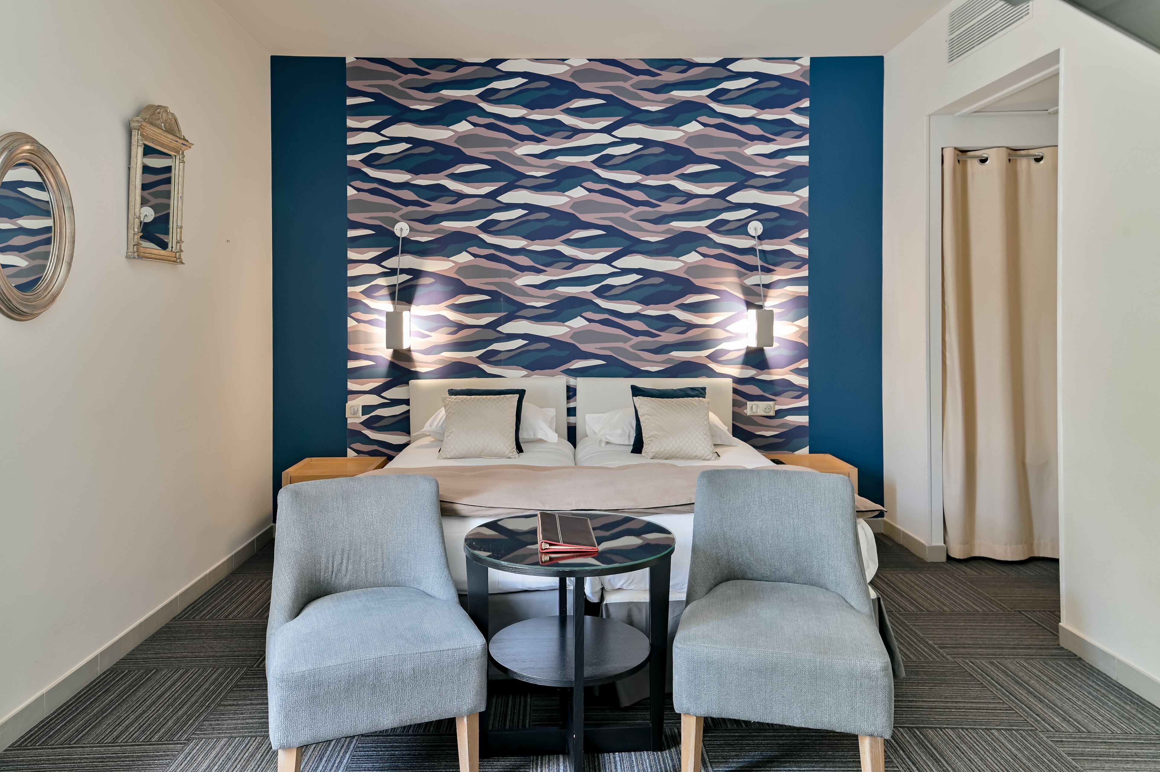 Una habitación de hotel con una cama, dos sillones, una mesita y un papel tapiz estampado.