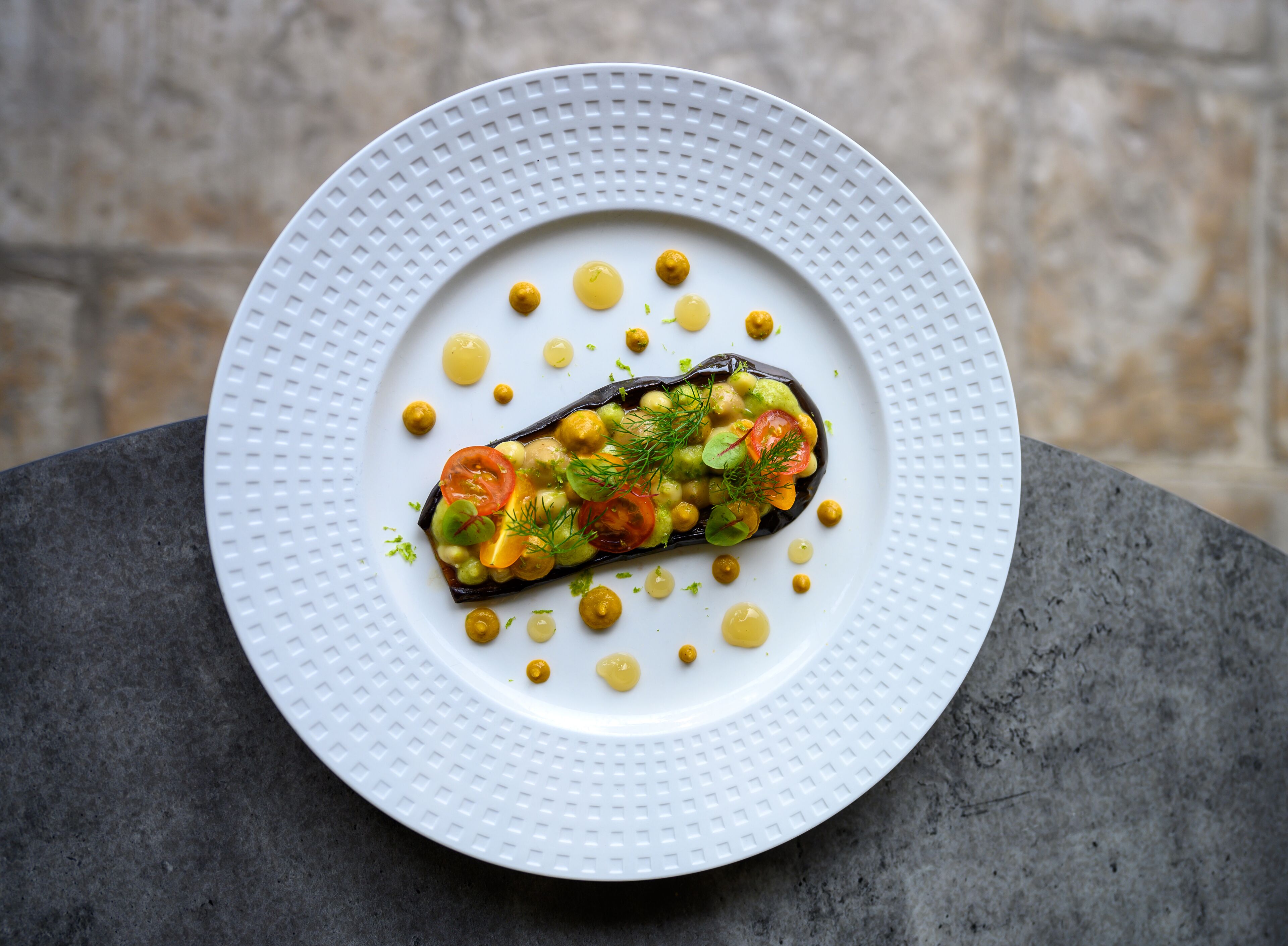 A white plate with a dish of stuffed eggplant, chickpeas, and vegetables, garnished with dill, on a dark surface.