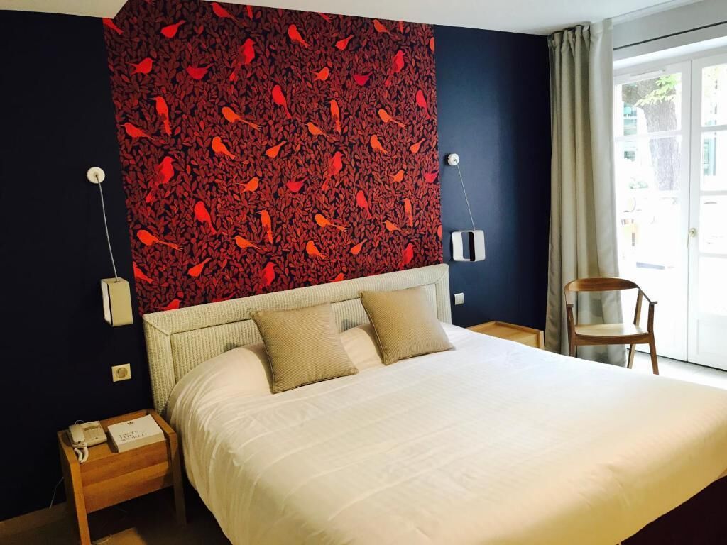 A hotel room with a large bed, a red patterned headboard, a wooden nightstand, a chair, and a window with curtains.
