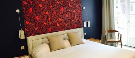 A hotel room with a large bed, a red patterned headboard, a wooden nightstand, a chair, and a window with curtains.