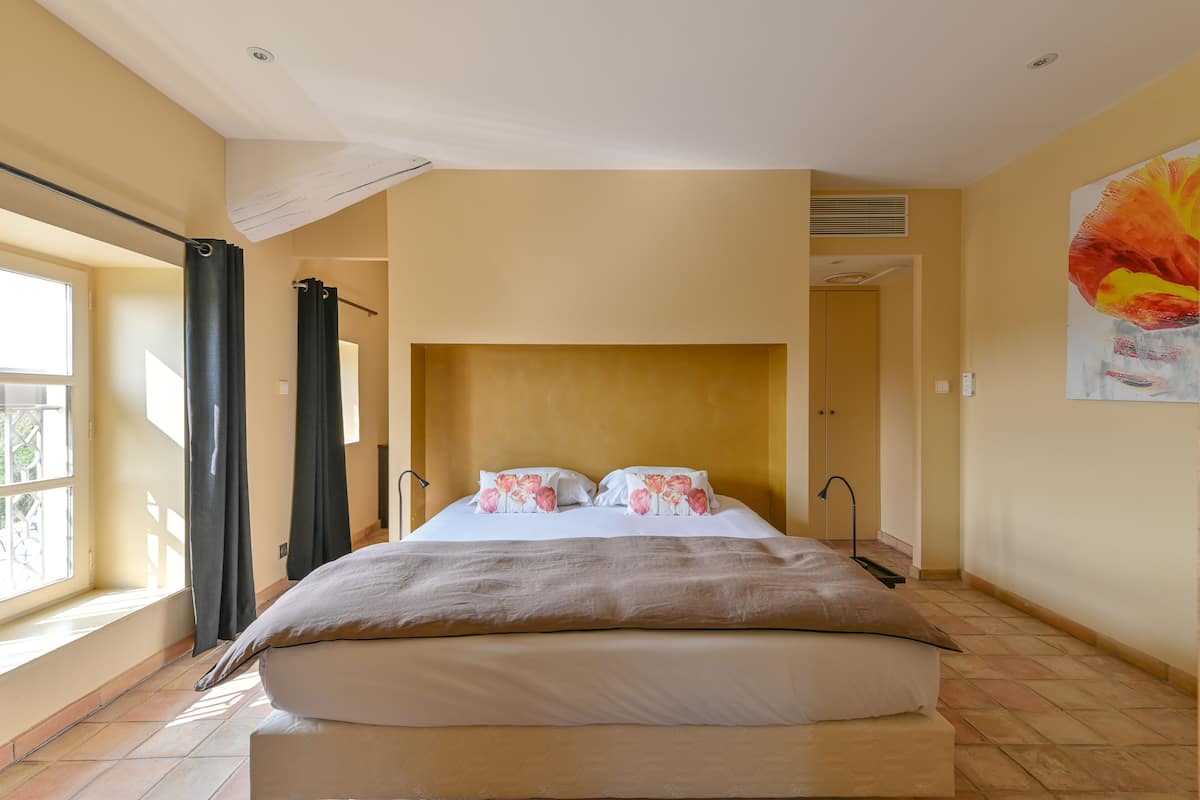 A bedroom with a large bed, a window with curtains, a painting on the wall, and a door leading to another room.