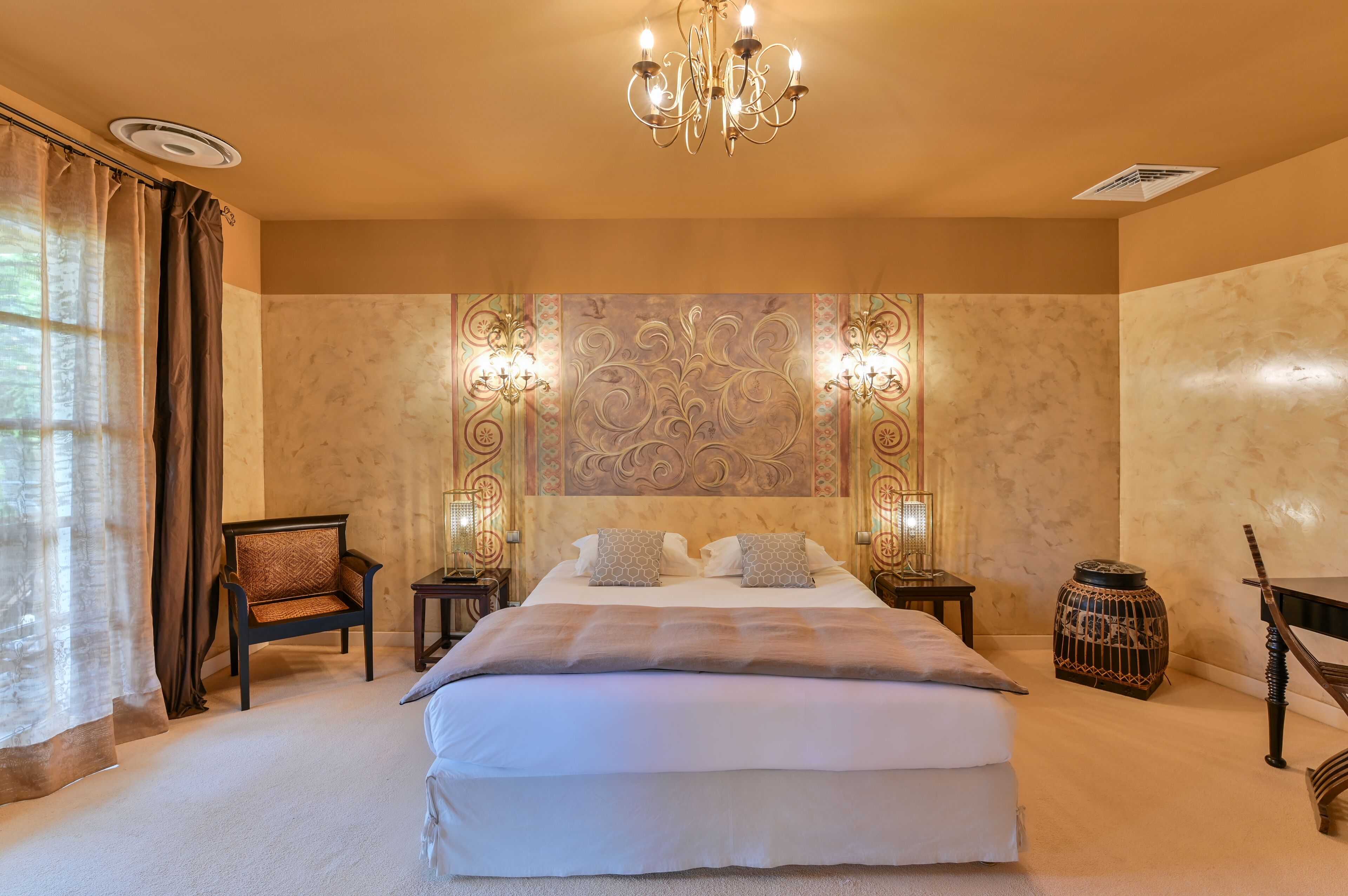 Ein geräumiges Schlafzimmer mit einem großen Bett, einer Chandelier, dekorativen Wandverkleidungen und einem kleinen Tisch mit einer Lampe.