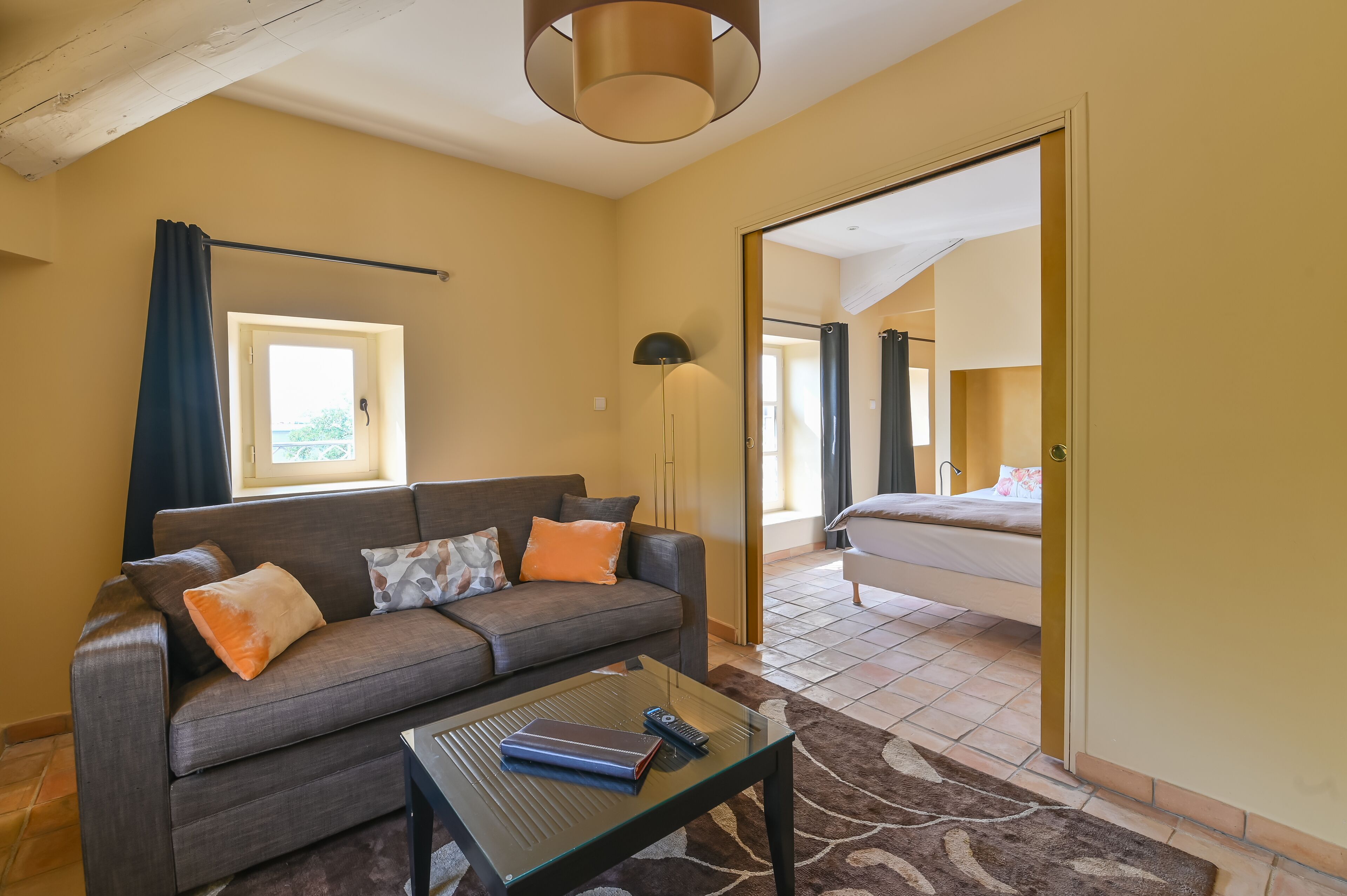 A modern living room with a sofa, coffee table, and a large mirror reflecting a bedroom with a bed.
