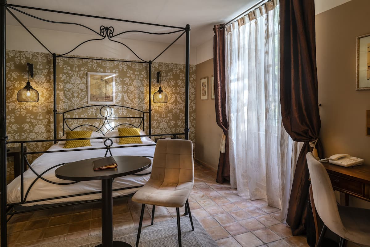A bedroom with a four-poster bed, a round table, a chair, and a desk with a telephone.