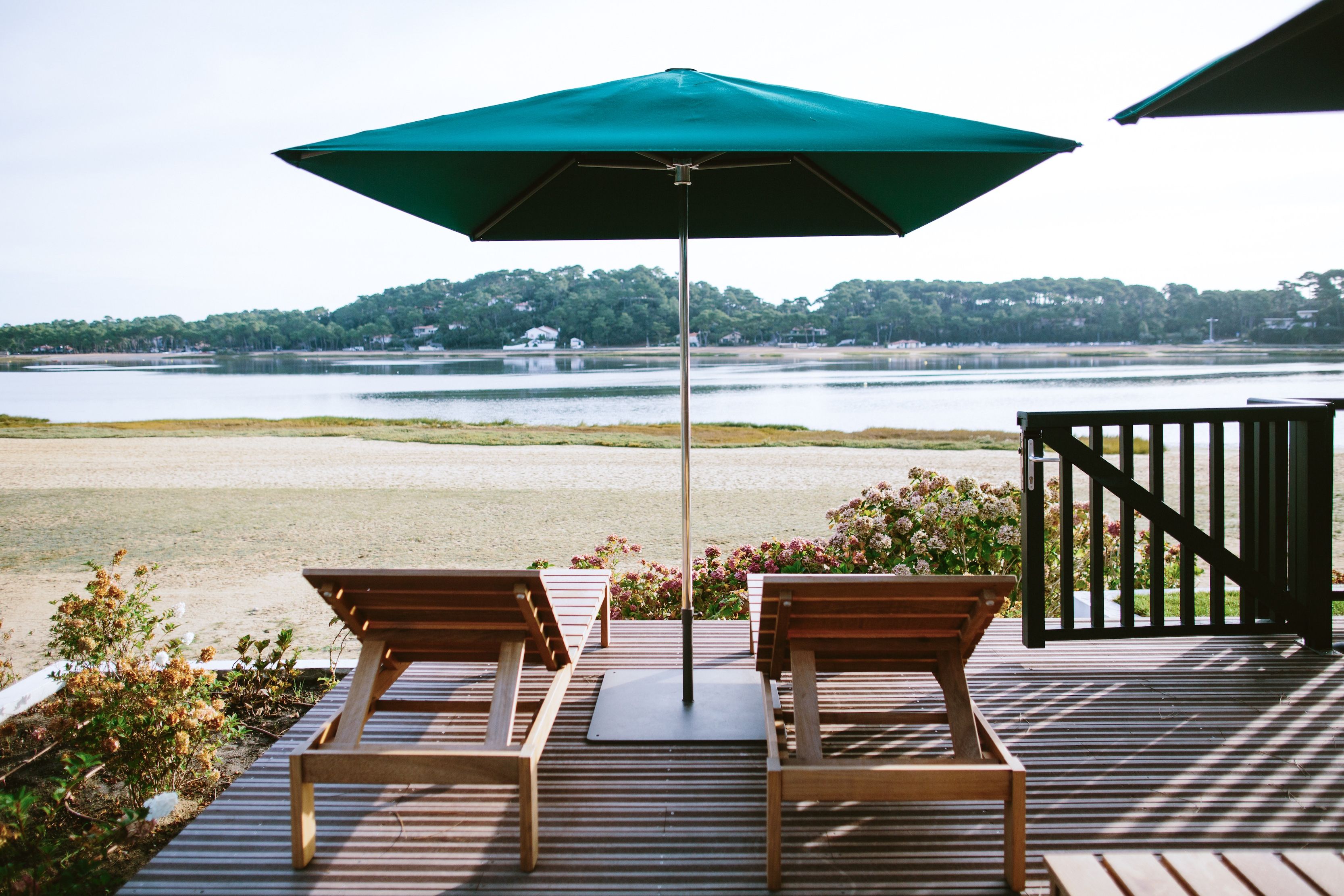 Terraza con dos sillones de madera y una sombrilla verde, con vista a un lago y una orilla boscosa.