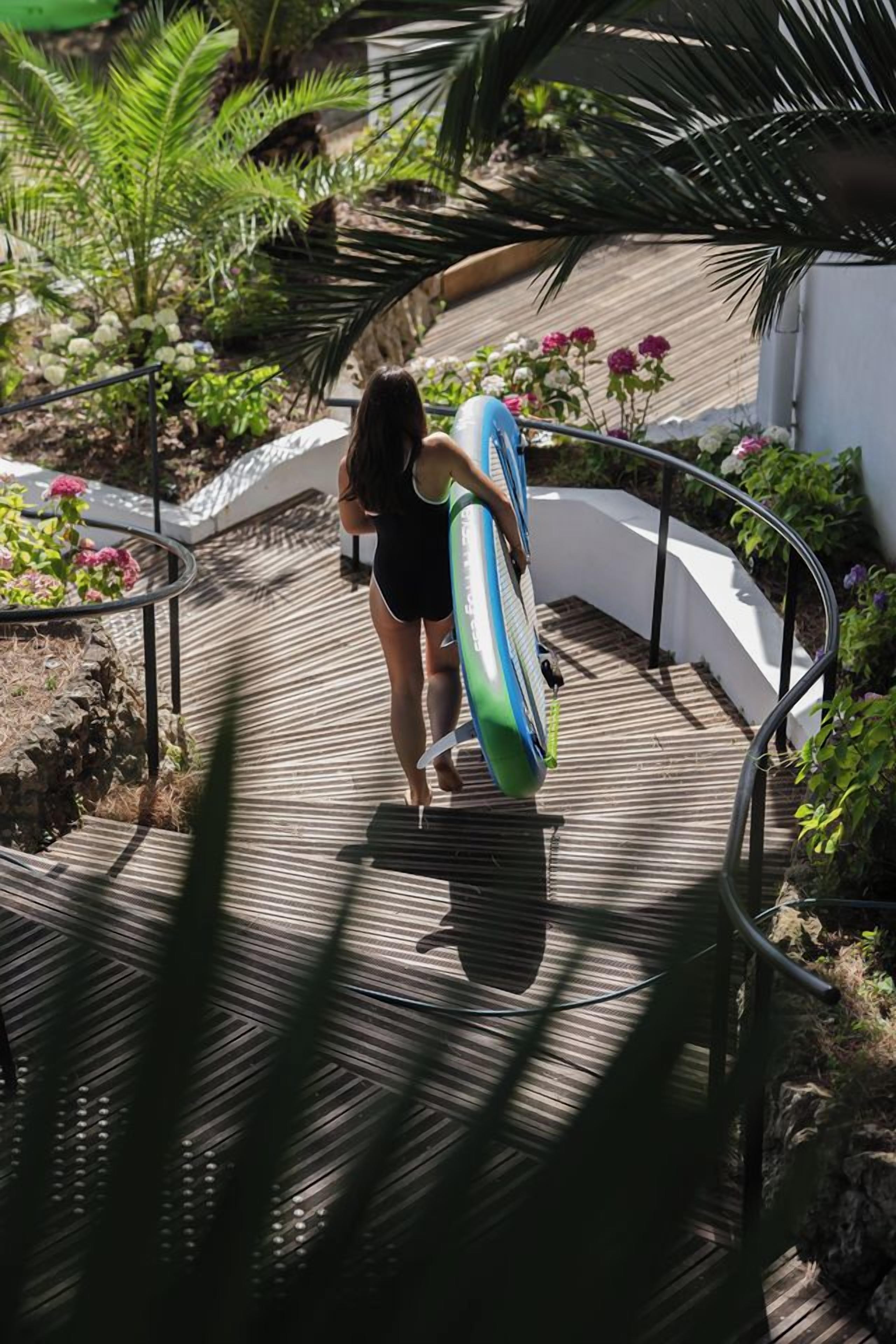A person walking on a wooden deck with a surfboard, surrounded by greenery and flowers.