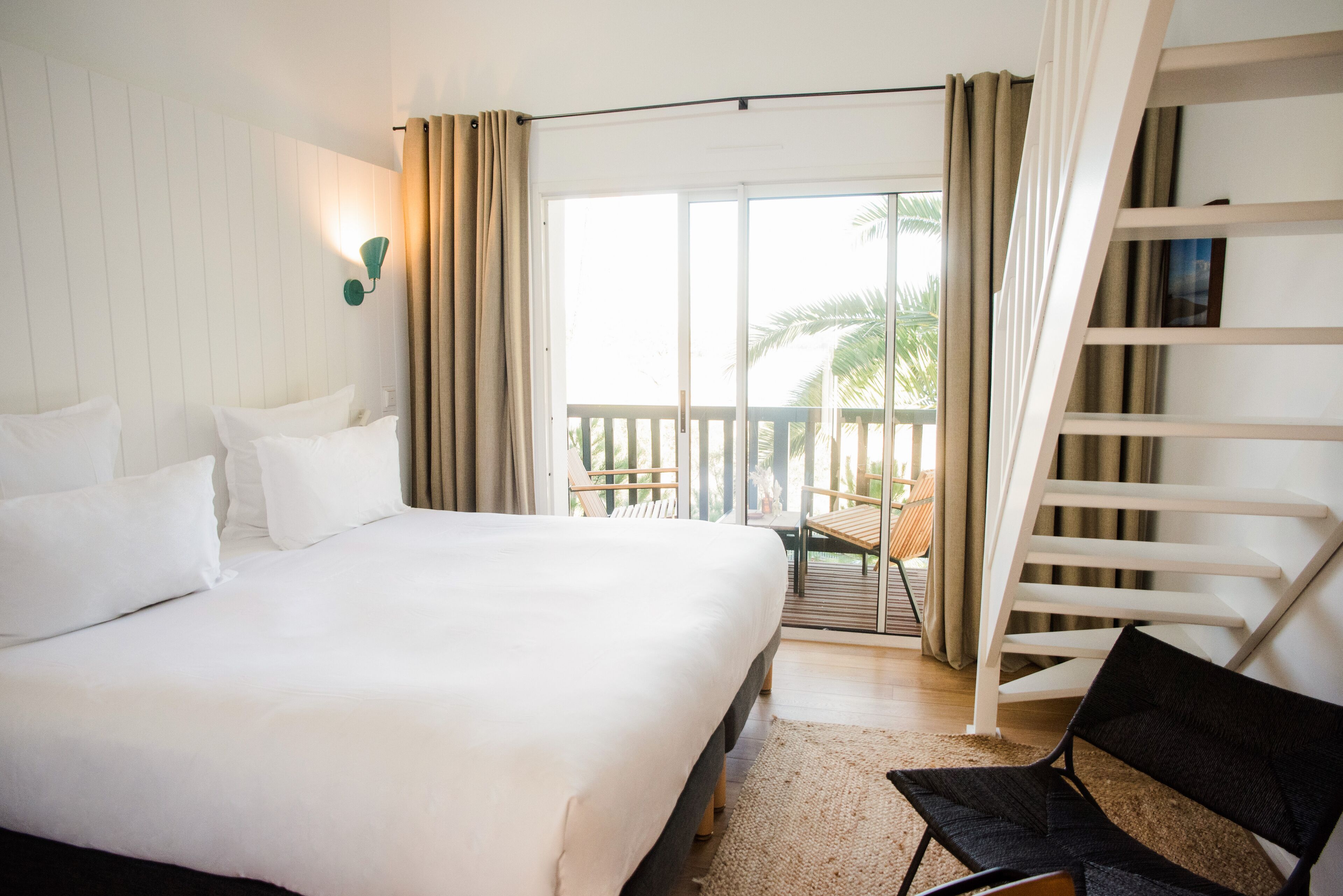 A bedroom with a large bed, a chair, a balcony with a view of palm trees, and a staircase.