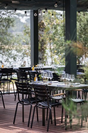 Un espace extérieur aménagé pour les repas, avec des tables et des chaises, une bouteille de vin et une vue sur les arbres.