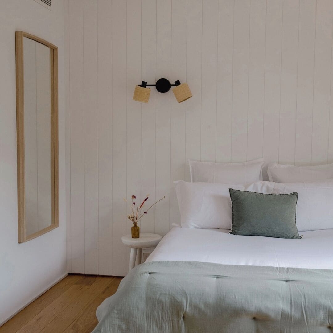 A bedroom with a bed, pillows, a bedside table, a mirror, and a wall-mounted light fixture.