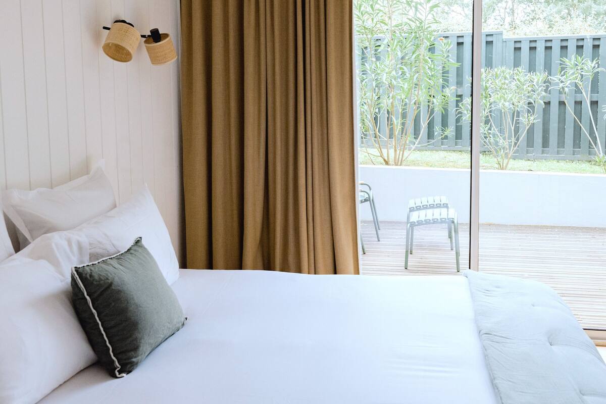 Ein Schlafzimmer mit einem Bett, weißen Kissen, einem braunen Vorhang und Blick auf einen Balkon mit Gartenmöbeln.