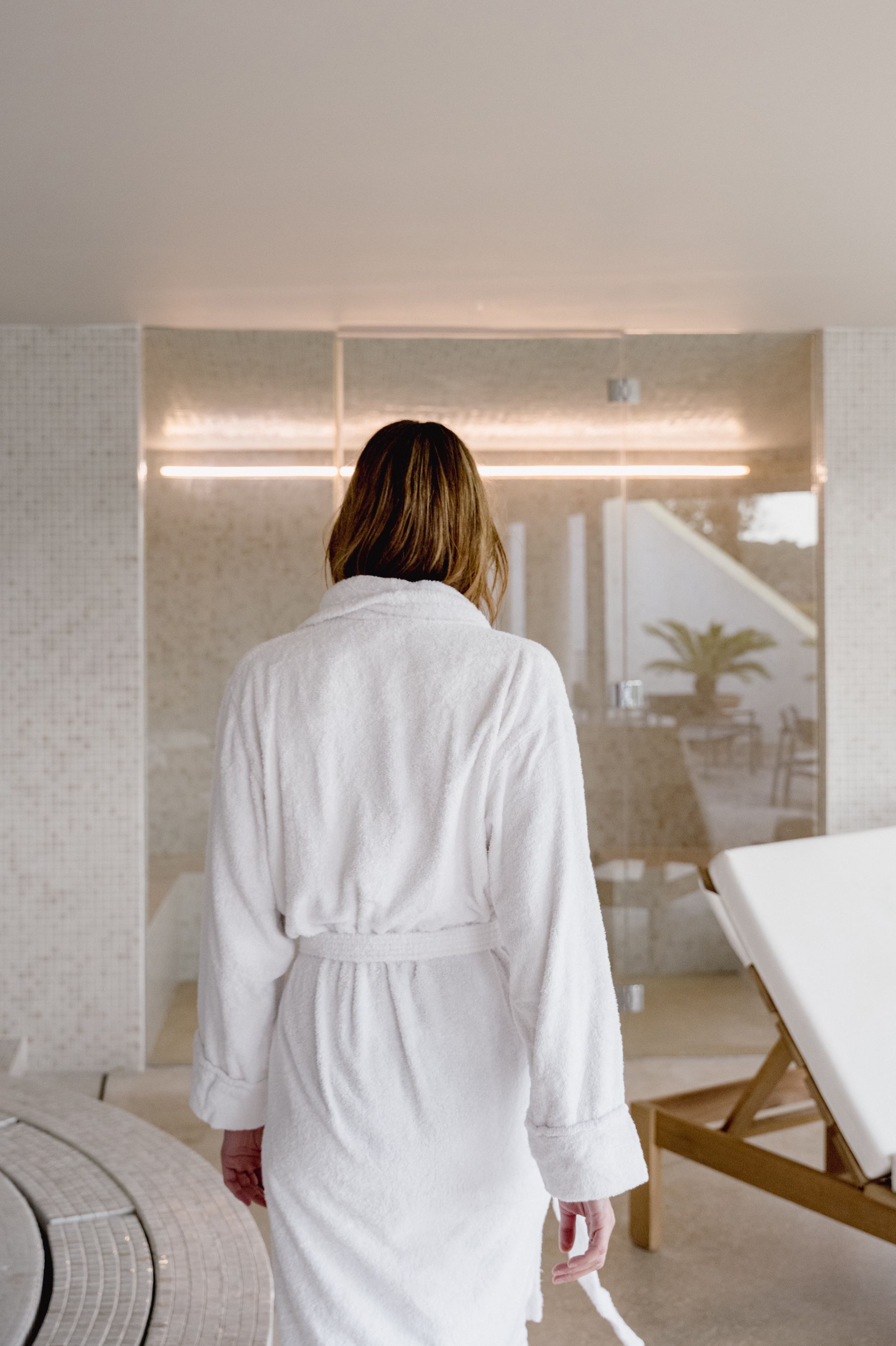 Una persona vestida con una bata blanca está de pie en una habitación con una cama, una camilla de masaje y vistas a un balcón con mobiliario exterior.