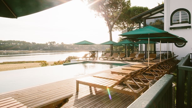 Un espace aménagé au bord de la piscine, avec des chaises longues en bois et des parasols, offrant une vue sur une étendue d’eau et un bâtiment doté de fenêtres en arc de cercle.