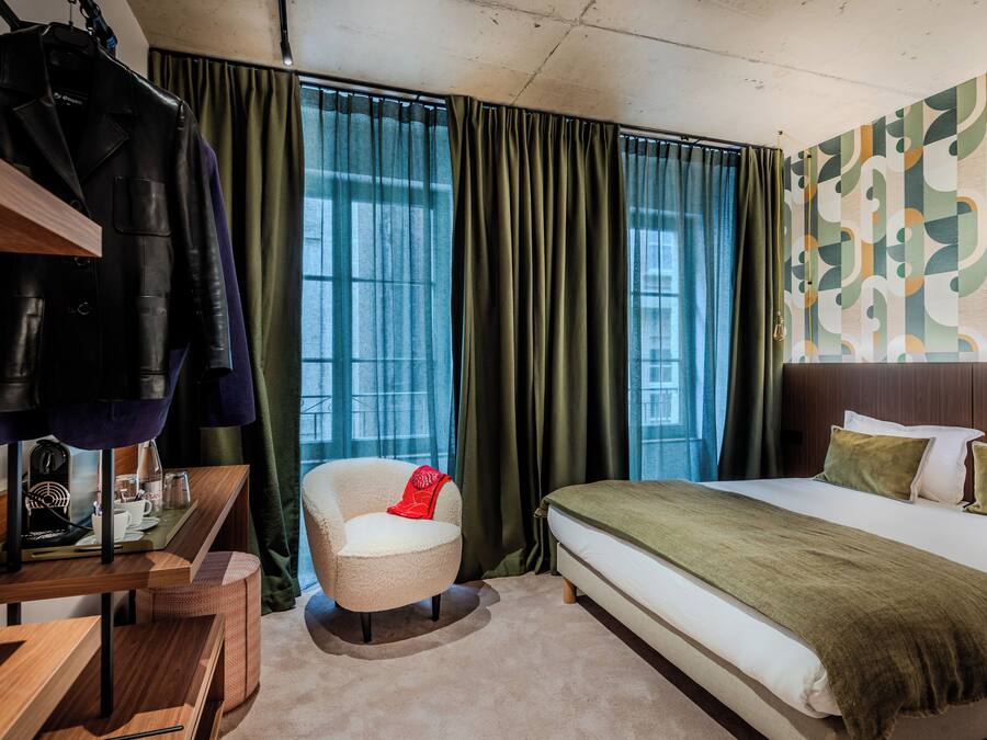 A hotel room with a bed, a chair, a desk, and a coat rack.