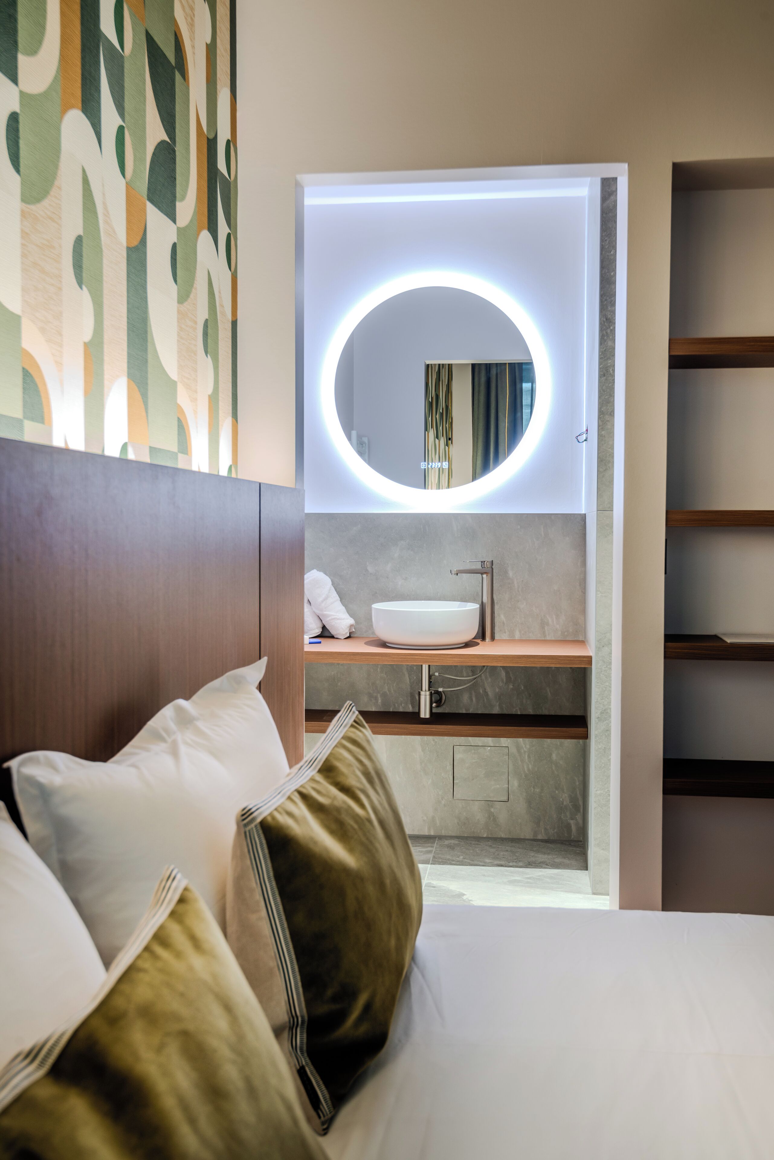 Una habitación de hotel moderna con una cama, una mesita de noche, un espejo redondo con luz, y un baño con lavabo y espejo.