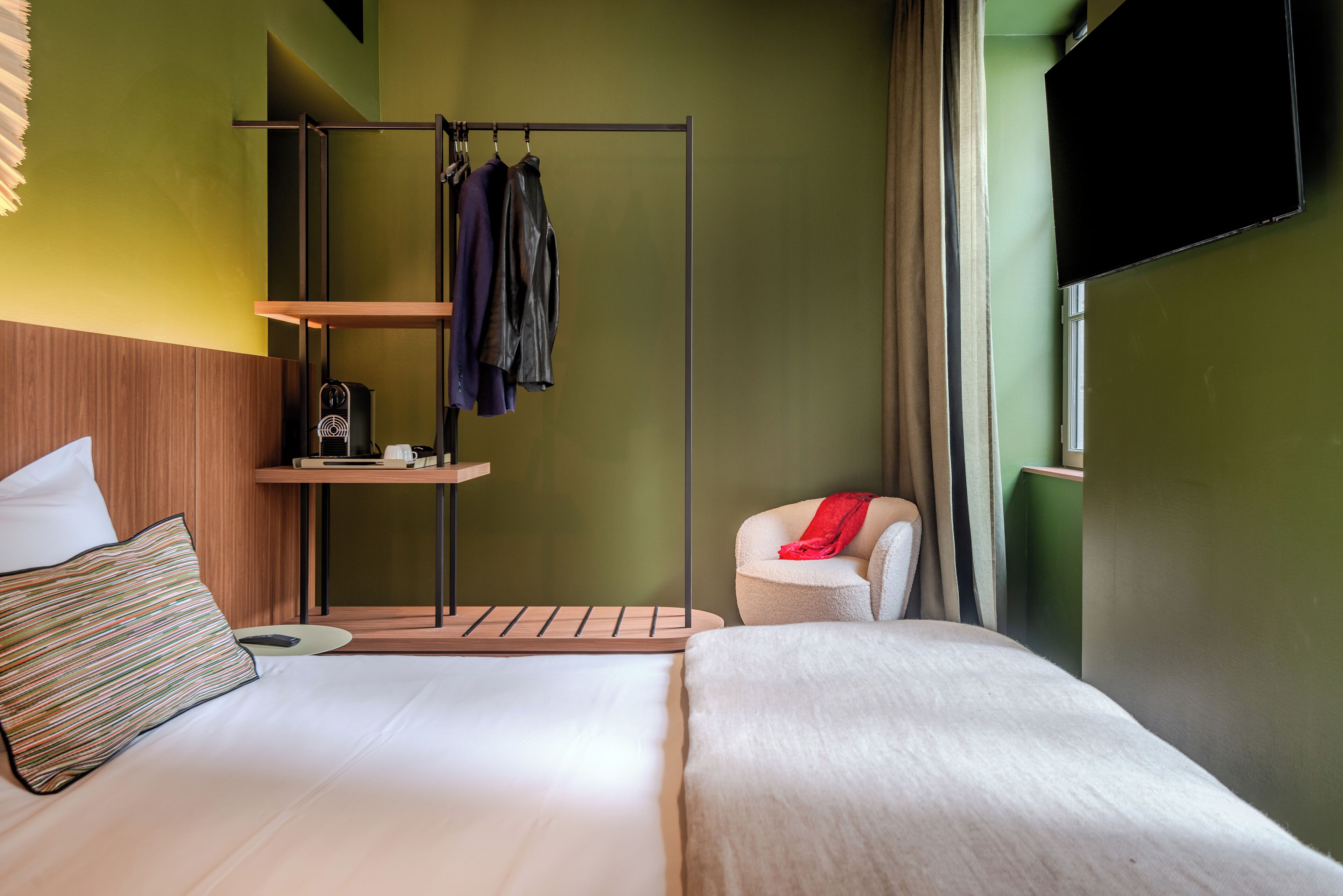 Ein Hotelzimmer mit Bett, einem kleinen Tisch mit Kaffeemaschine, einem Kleiderständer und einem an der Wand befestigten Fernseher.