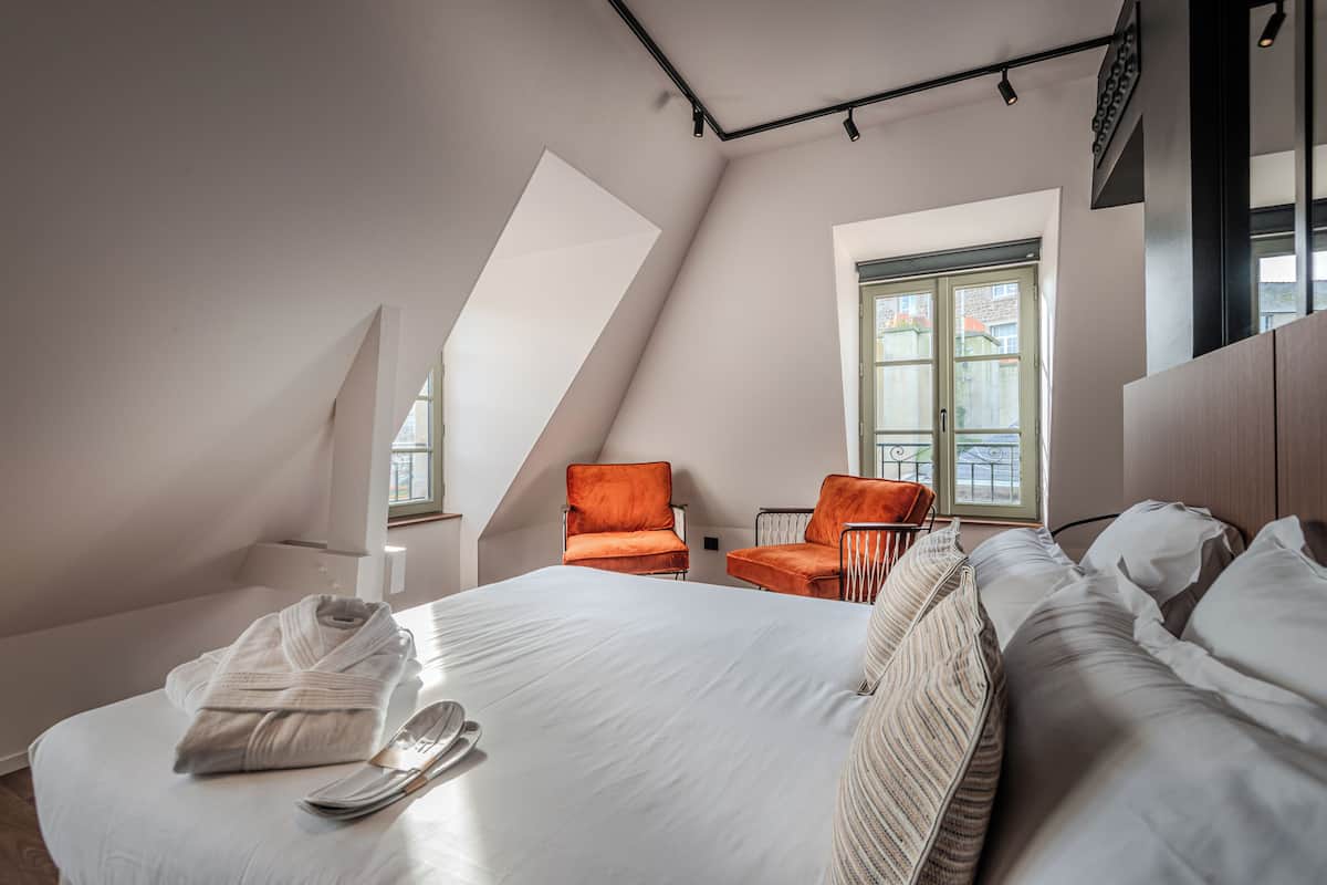 A modern hotel room with a large bed, two orange chairs, a window with a view, and a sloped ceiling.
