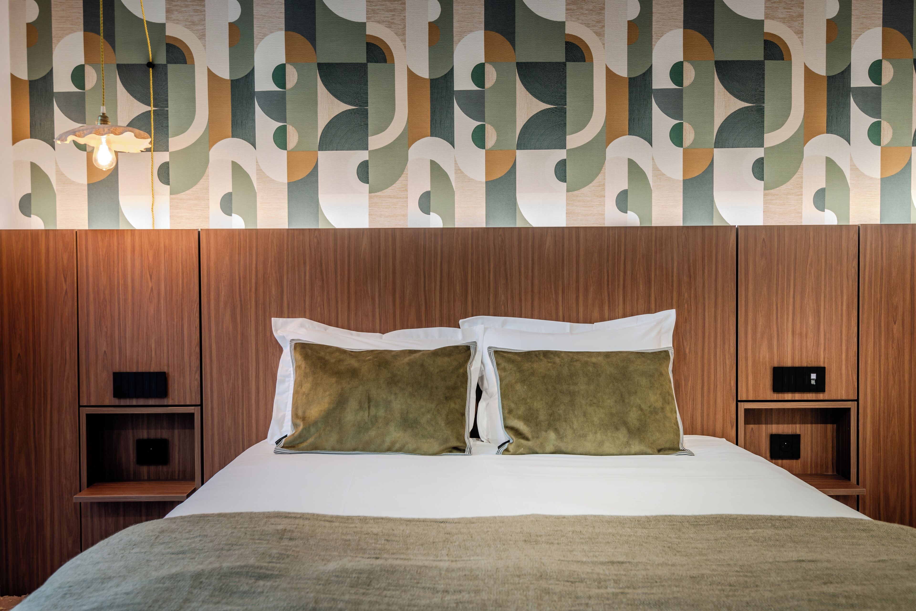 Habitación de hotel con cabecera de madera, dos mesitas de noche, cama con ropa blanca y pared con estampado geométrico.