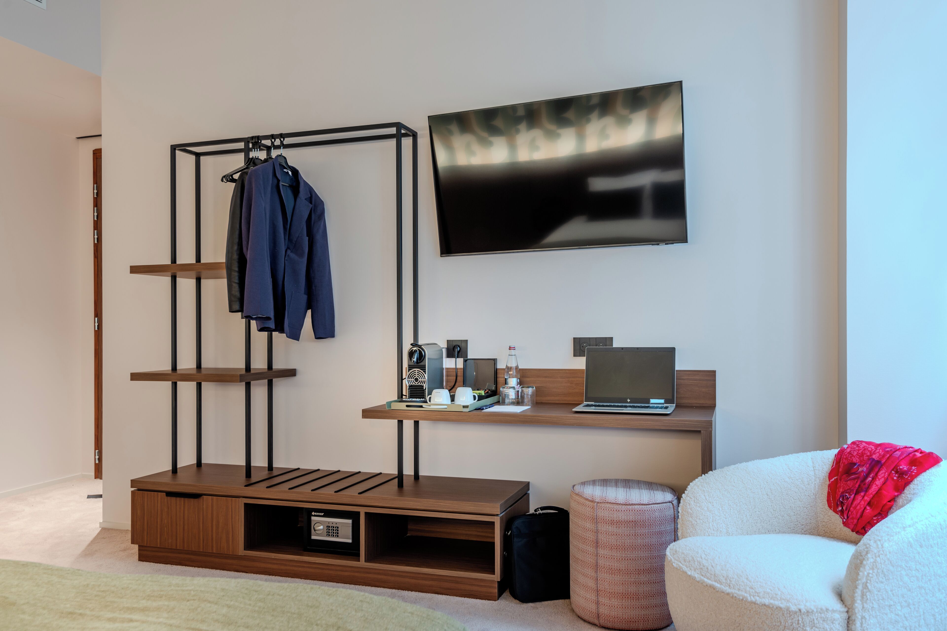 Een moderne kamer met een houten bureau, een wandgemonteerde televisie, een kledingrek en een gezellige zithoek.