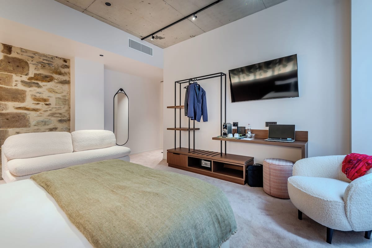 A modern bedroom with a stone wall, a hanging clothes rack, a bed with a green blanket, a white sofa, a small ottoman, and a TV on the wall.