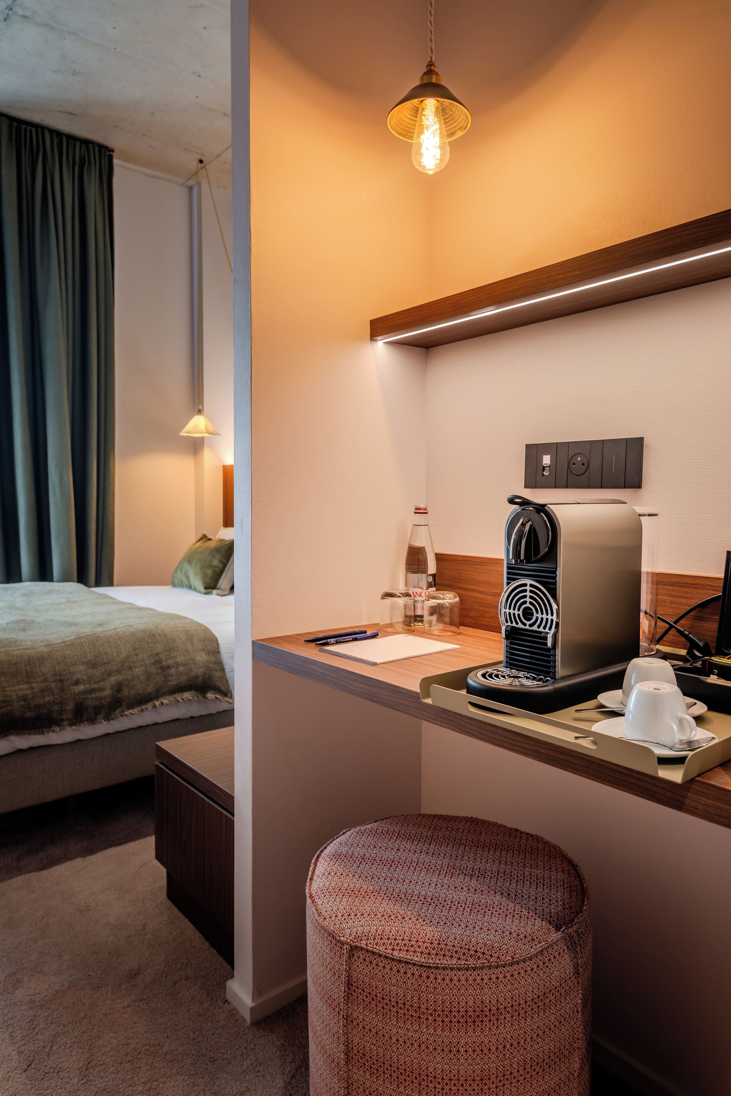 A hotel room with a bed, a small kitchenette with a coffee maker, a bottle of water, and a cup.