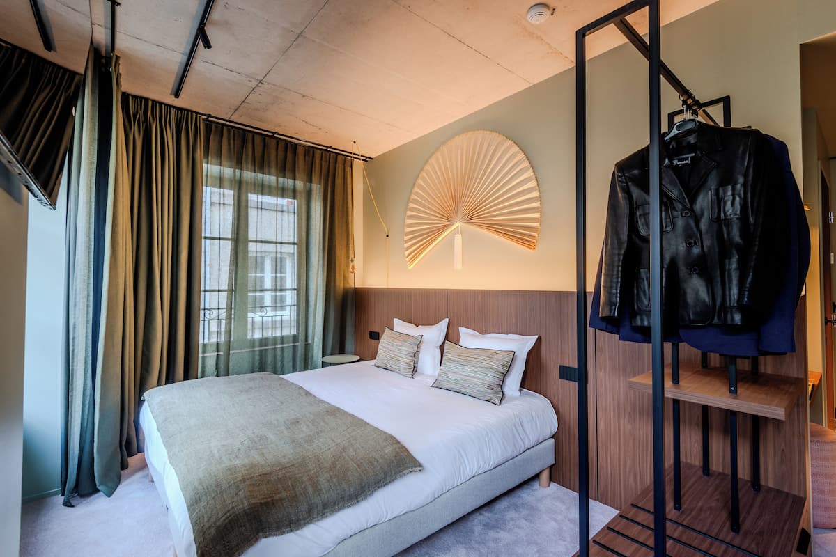 A modern hotel room with a large bed, a wooden headboard, a hanging coat rack, and a wall-mounted fan.