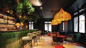 Een bar met een groen aanrecht, meerdere barkrukken en hangplanten. Er zijn verschillende tafels en stoelen voor het zitten.