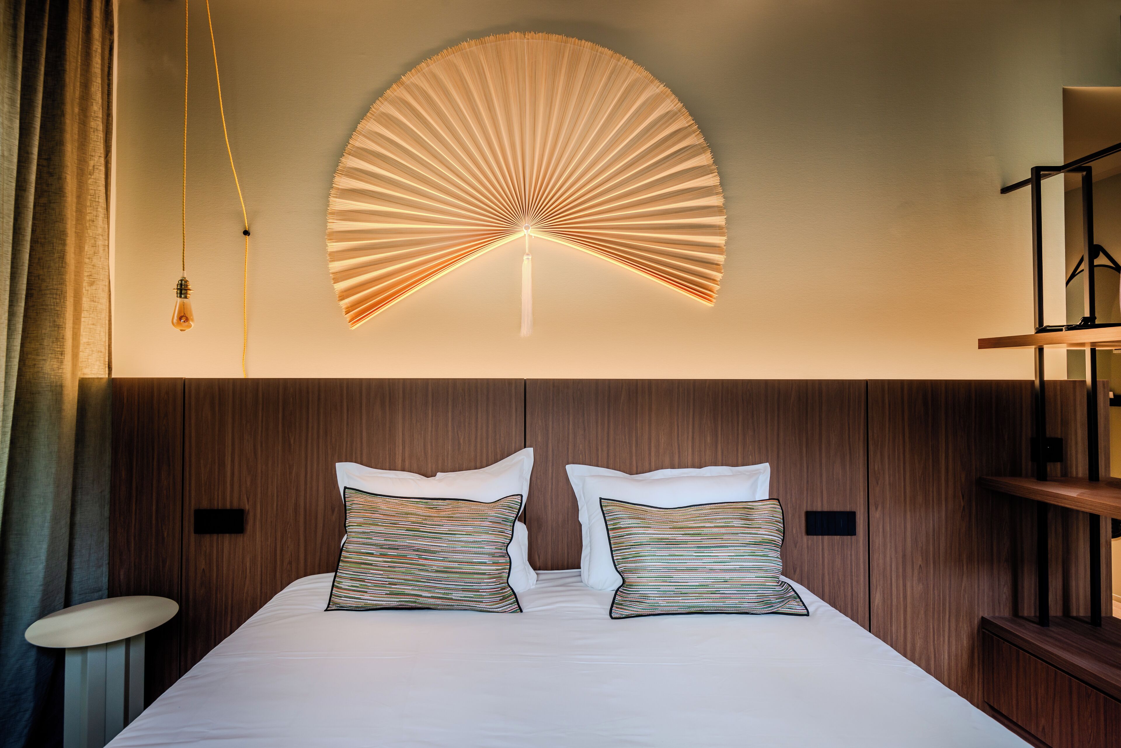 Et moderne hotelværelse med en stor seng, to puder, en vægmonteret ventilator og et natbord.