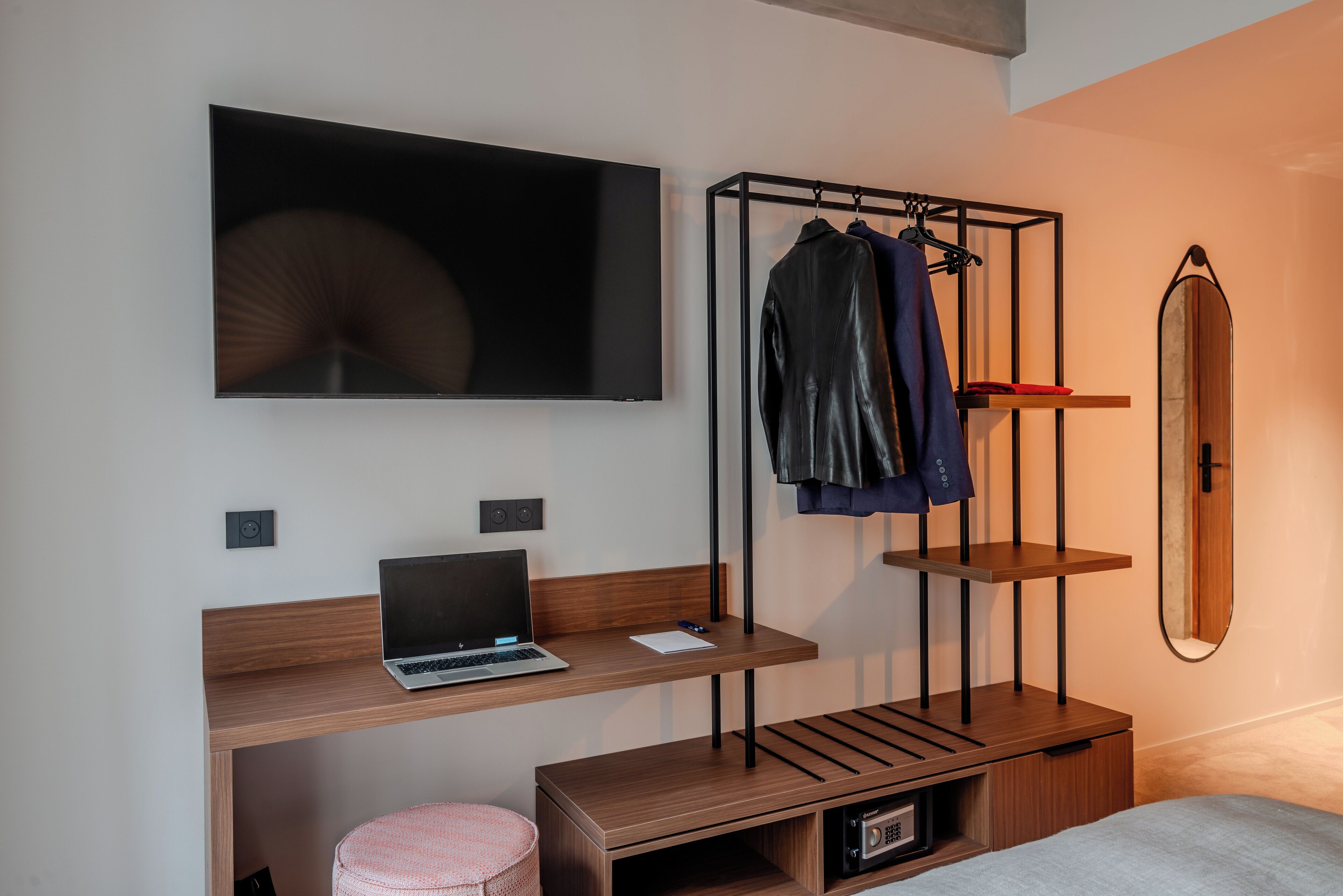 Une chambre moderne avec un bureau en bois, un ordinateur portable, un porte-manteau suspendu, un miroir et un lit.