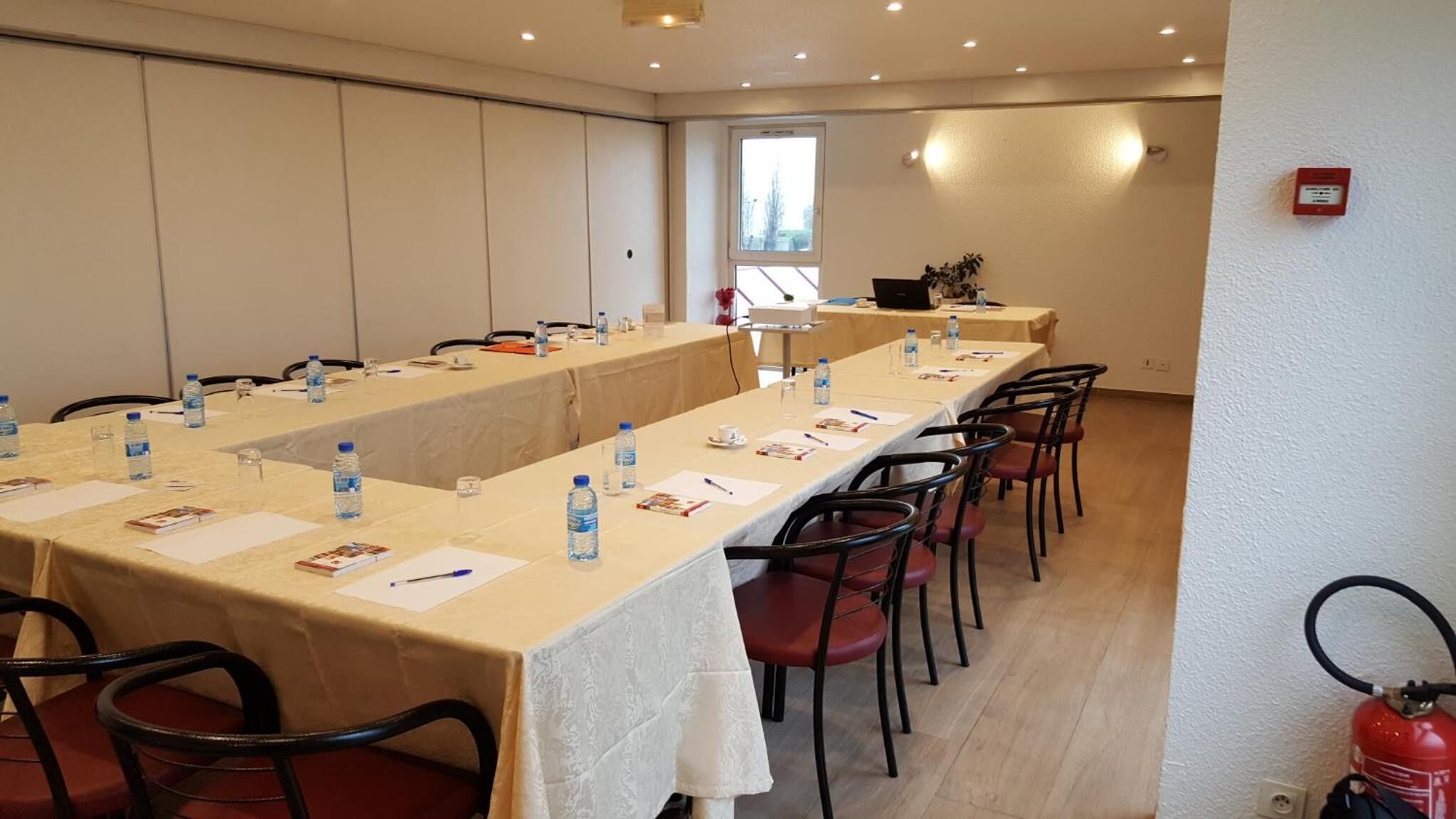 Ein Konferenzraum mit langen Tischen, Stühlen, Wasserflaschen und einem Feuerlöscher.