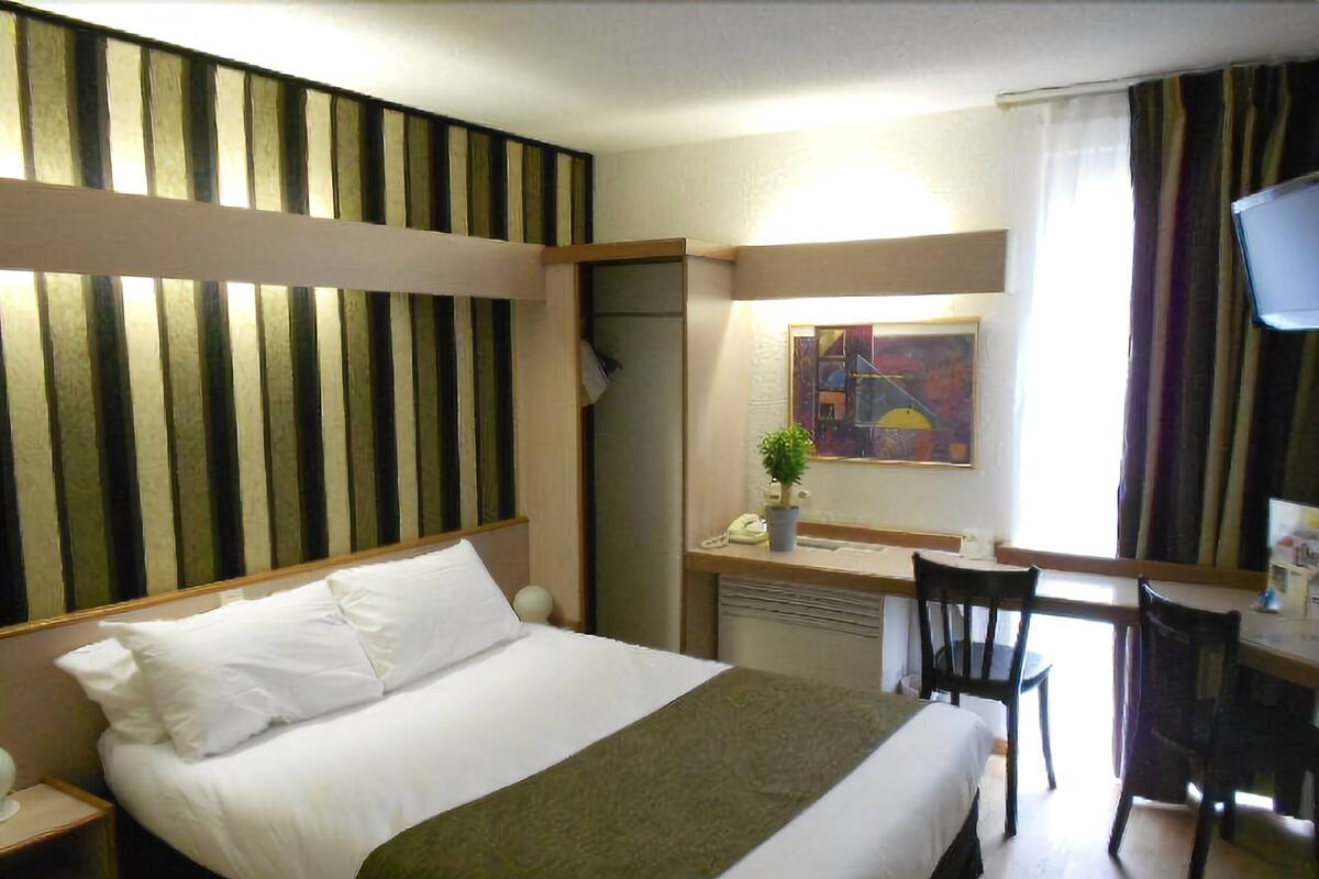 Habitación de hotel con cama, escritorio, silla, televisor y ventana con cortinas.