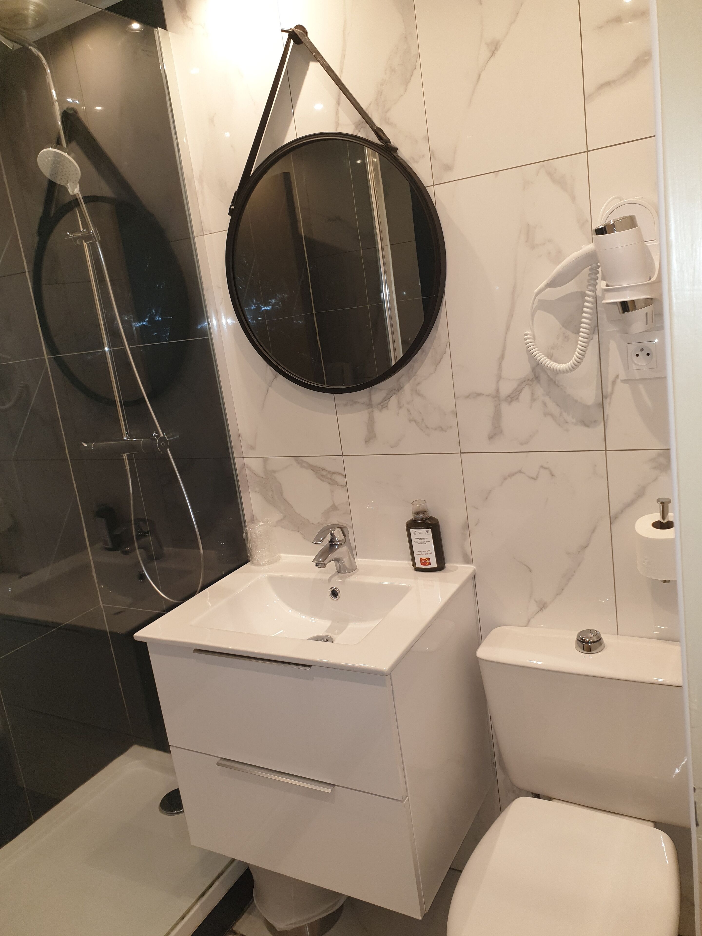 Une salle de bain moderne avec un lavabo blanc, un miroir rond, une douchette à main et des toilettes.