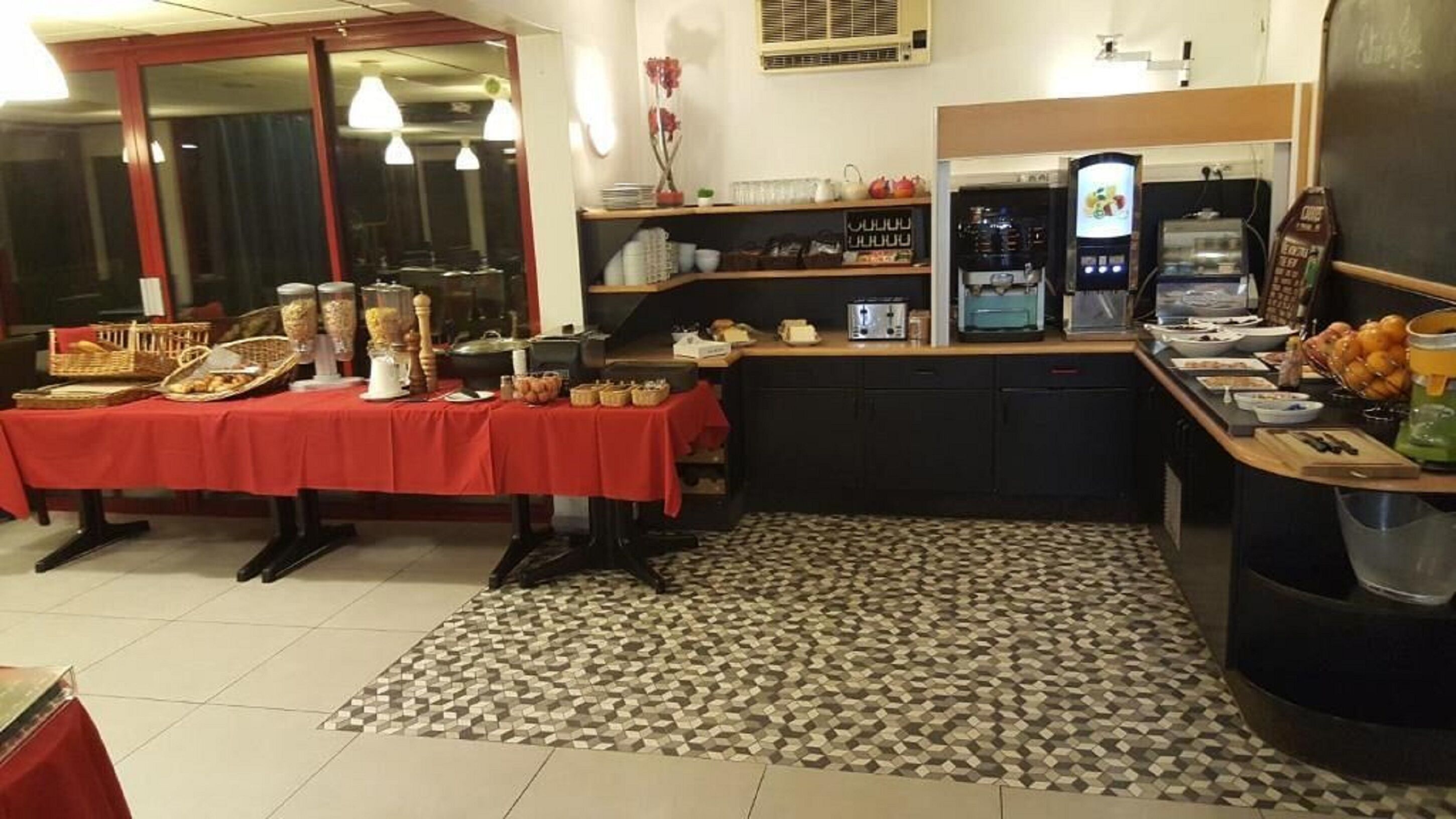 Una cocina con una mesa roja, diversos alimentos, una cafetera y un refrigerador.
