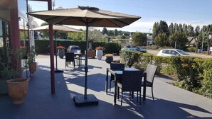 Un patio al aire libre con una mesa y sillas bajo un paraguas grande, rodeado de plantas en macetas y vista a la calle.