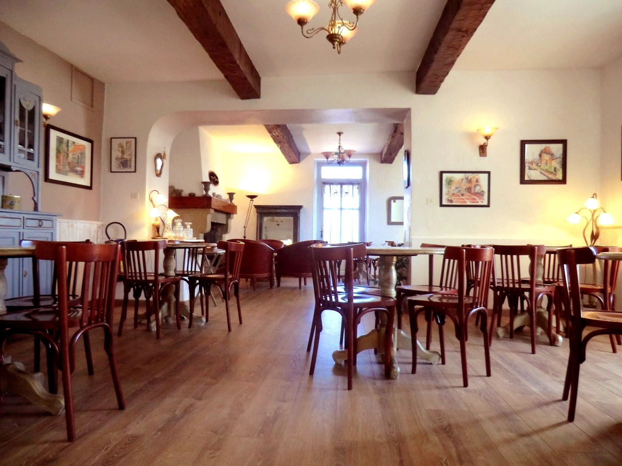 Un vaste espace dédié à la salle à manger, doté d’un plancher en bois et de nombreuses tables et chaises en bois. Les murs sont ornés de tableaux encadrés et un lustre est suspendu au plafond.