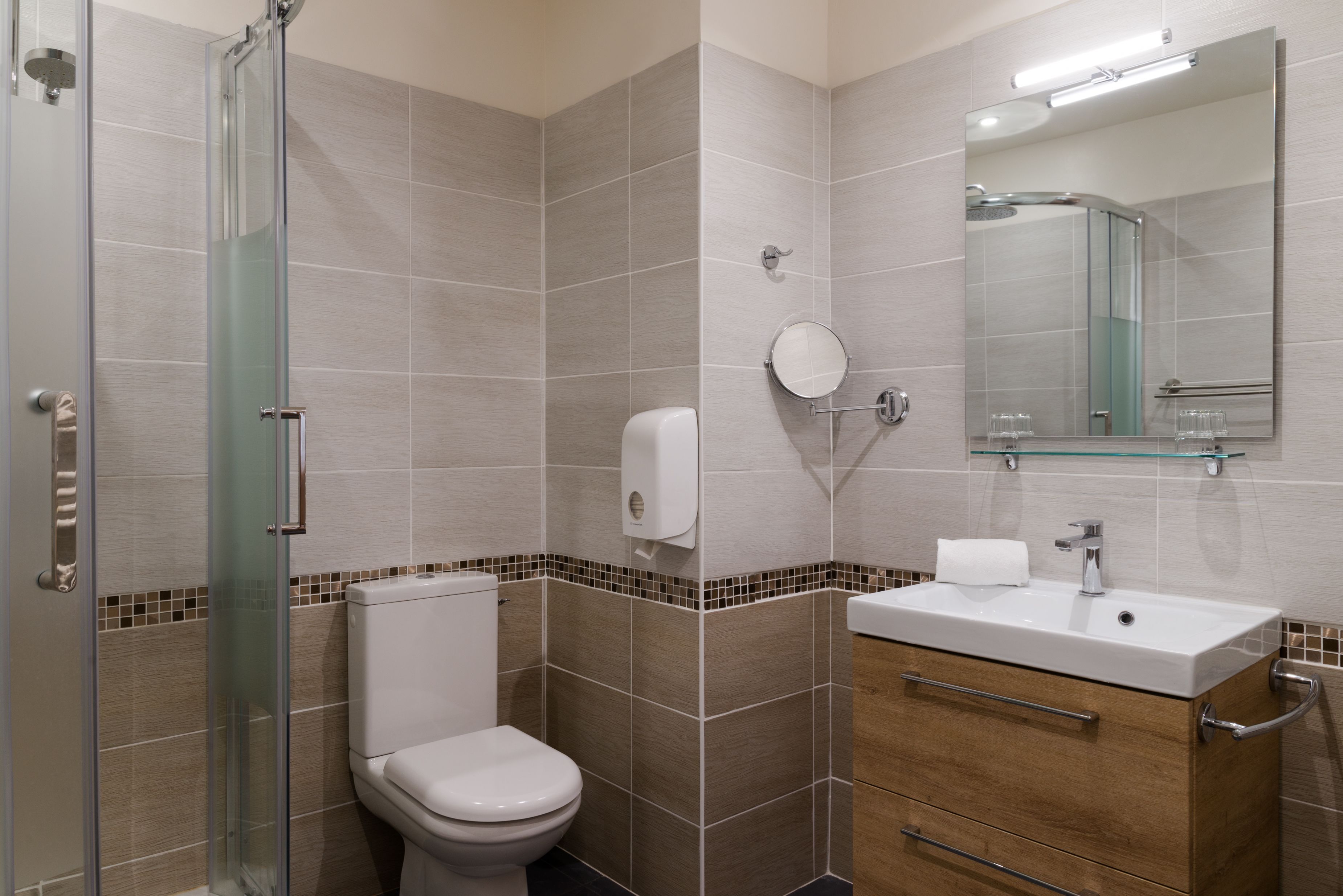 A modern bathroom with a glass shower enclosure, a toilet, a sink with a mirror, and a towel dispenser.