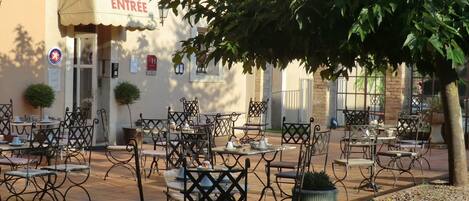 Un área de café al aire libre con mesas y sillas, la entrada de un edificio con un letrero, y un árbol que proporciona sombra.
