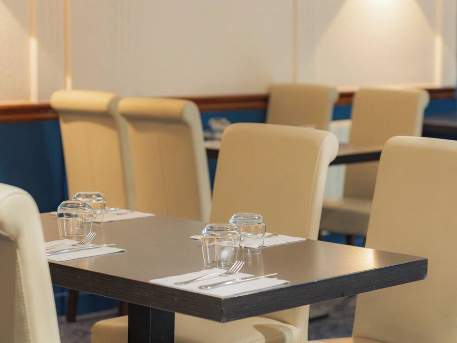 Área de refeições de hotel com mesas para quatro pessoas, cadeiras e luminária fixada na parede.