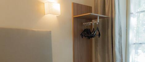 A hotel room with a bed, a bedside table, a hanging coat rack, and a window with curtains.