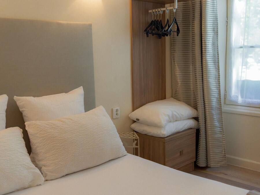 Quarto de hotel com cama, mesa de cabeceira, cabideiro e janela com cortinas.